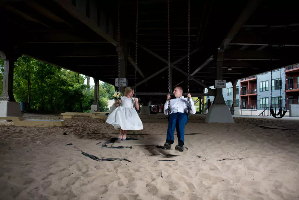 Katie and Zack enjoy swinging together at Swing Park beneath the Holton Street Bridge in Milwaukee during their wedding reception. The urban park setting with colorful swings adds a playful and joyful vibe to the celebration.