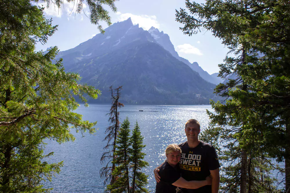 Katie and Zack stand smiling by the shore of Jenny Lake in Grand Teton National Park, with calm water reflecting the surrounding pine trees and mountain peaks. The peaceful lake scene highlights the natural beauty of the Wyoming park.