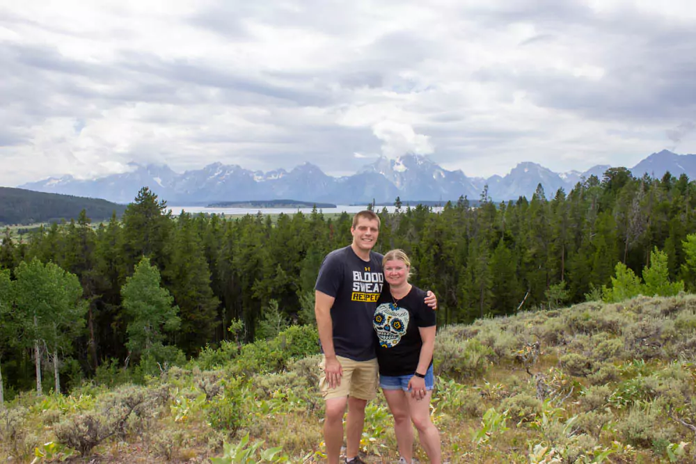 Katie and Zack hike along a scenic trail in Grand Teton National Park, surrounded by tall pine trees and towering mountain peaks under a clear blue sky. The photo captures their outdoor adventure in the rugged Wyoming wilderness.