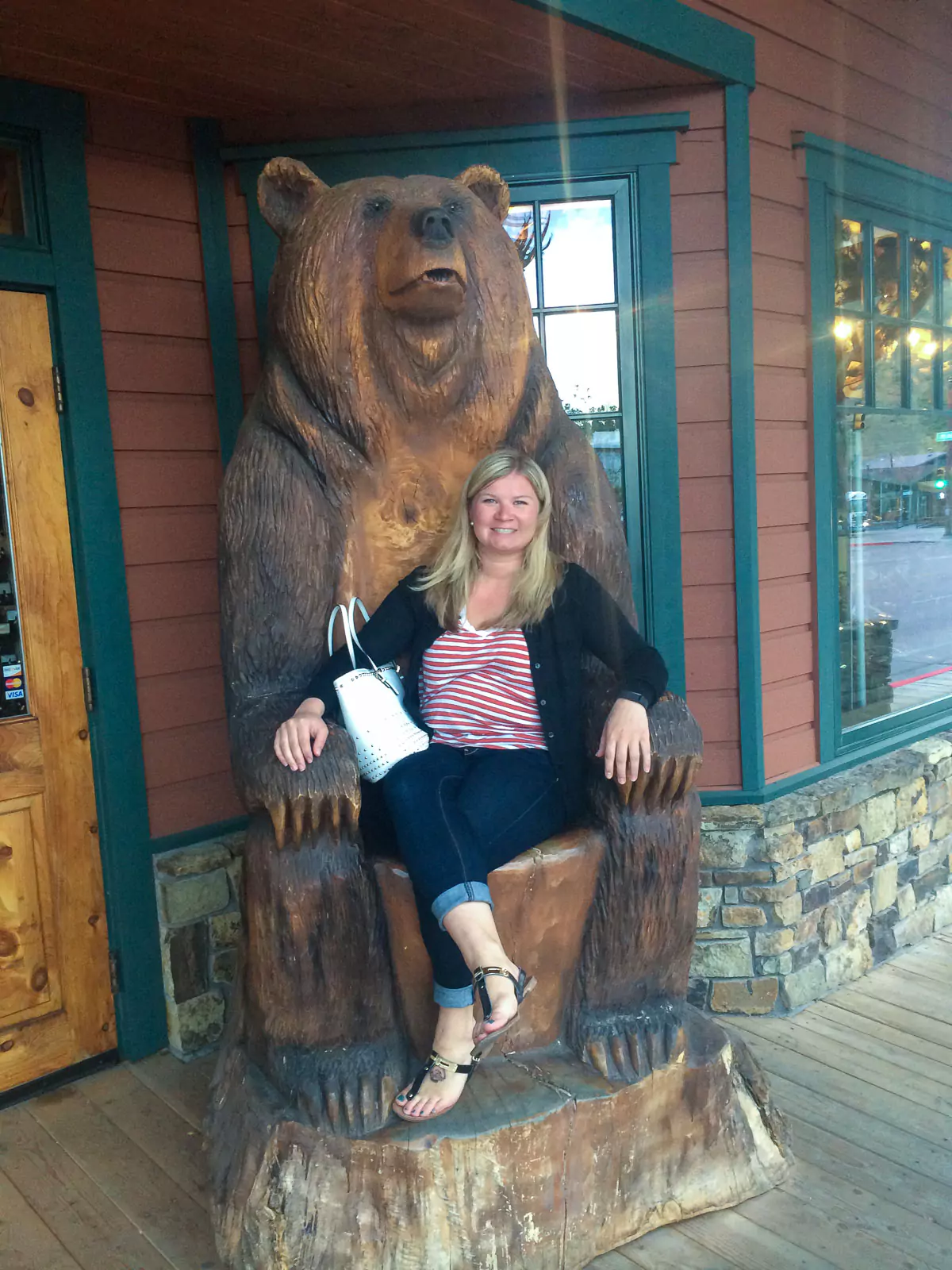 Katie sits smiling on a large wooden bear carving in Jackson Hole, Wyoming, surrounded by rustic outdoor scenery. The detailed sculpture captures the western spirit of the area.