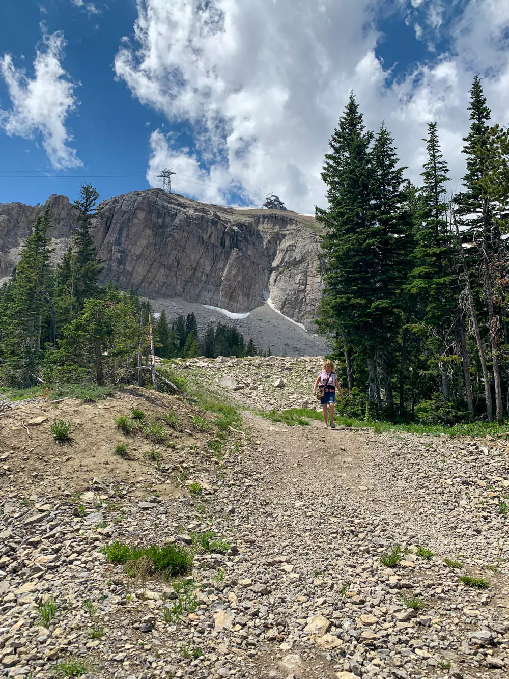 Barb hikes along the Cirque Trail at Jackson Hole Mountain Resort, surrounded by green trees and mountainous terrain under a clear blue sky. The trail offers scenic views and outdoor adventure in the Wyoming wilderness.