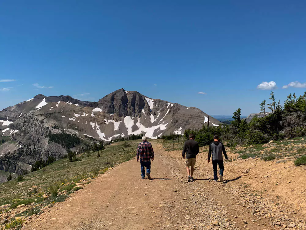 Dad, Zack, and Nick walk together along a mountain trail at Jackson Hole Mountain Resort, surrounded by pine trees and rugged peaks under a clear sky. The group enjoys a scenic hike in the Wyoming wilderness.