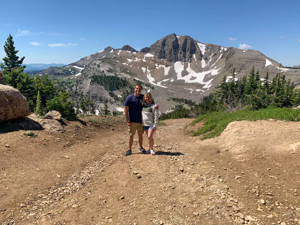Katie and Zack hike along a mountain trail near the summit of Jackson Hole Mountain Resort, surrounded by pine trees and rugged peaks under a clear blue sky. The scenic view highlights their outdoor adventure in Wyoming’s wilderness.