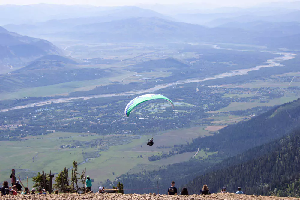 A man paraglides over the rugged mountain landscape of Jackson Hole Mountain Resort in Wyoming, soaring high above pine-covered slopes under a clear blue sky. The adventurous scene highlights the thrill of aerial sports in the scenic wilderness.