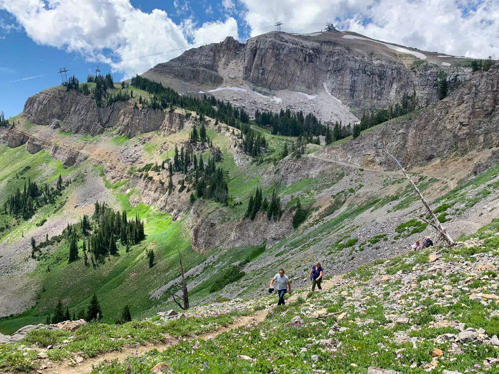 Nick and Mary hike along a scenic mountain trail at Jackson Hole Mountain Resort, surrounded by pine trees and rugged peaks under a clear blue sky. The outdoor adventure captures the natural beauty of the Wyoming landscape.