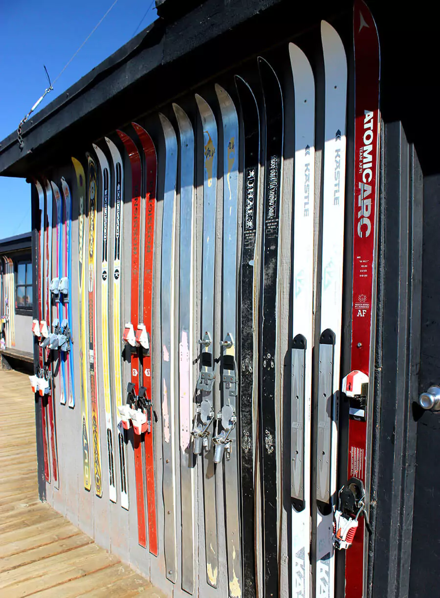 Several pairs of vintage skis mounted as decorative elements on the exterior wall of Corbet’s Cabin at Jackson Hole Mountain Resort. The rustic wooden cabin is set against a snowy mountain backdrop.