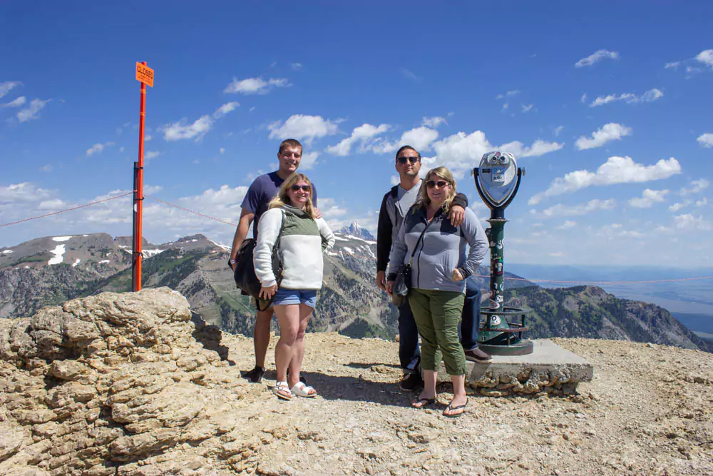 Zack, Katie, Mary, and Nick stand together at the summit of a mountain at Jackson Hole Mountain Resort, surrounded by panoramic views of rugged peaks and pine forests under a clear sky. The group smiles, capturing a memorable moment in the scenic Wyoming wilderness.
