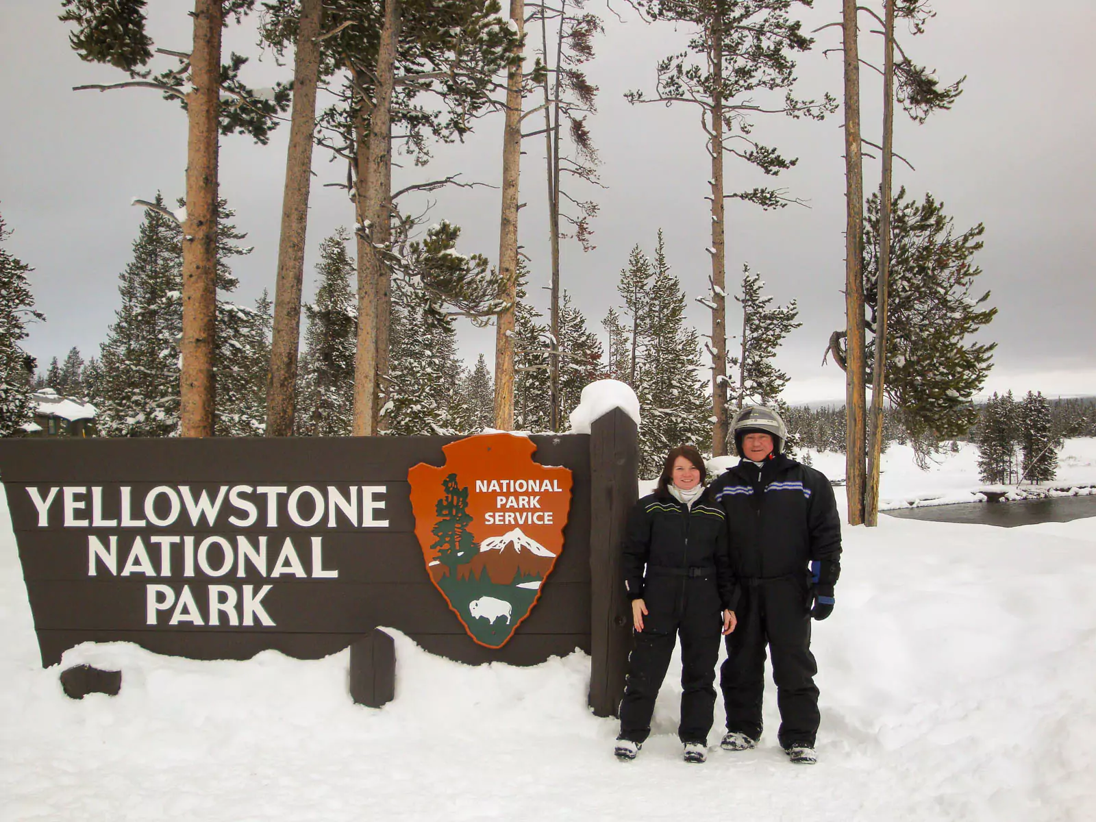 Katie and her dad pose on a snowmobile beside the Yellowstone National Park entrance sign, partially covered by snow reaching up to the bottom of the sign. The snowy landscape highlights a winter visit to the park.