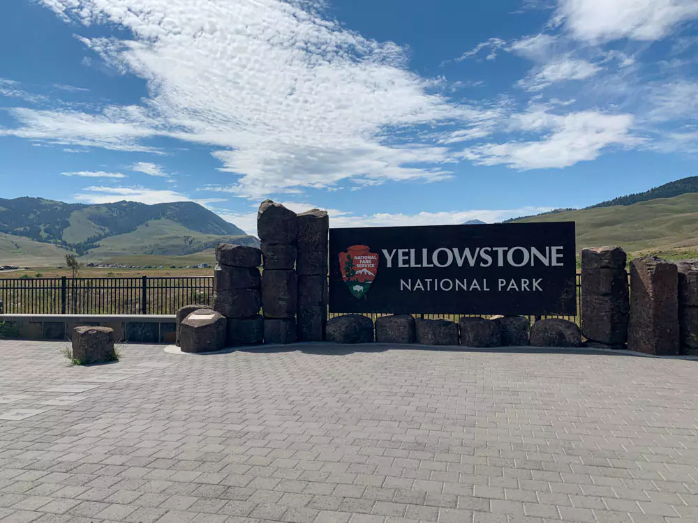 A large wooden entrance sign for Yellowstone National Park located in Montana, set against a backdrop of pine trees and a clear sky. The sign welcomes visitors to the expansive national park.